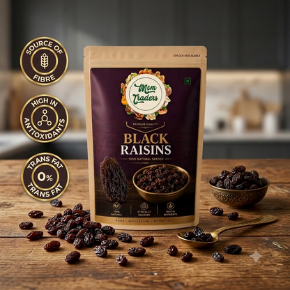 A stand-up pouch of Mom Leaders Black Raisins on a rustic wooden table, with loose raisins scattered around and a small bowl of raisins to the right. Three gold circular badges on the left highlight source of fibre, high in antioxidants, and trans fat free.