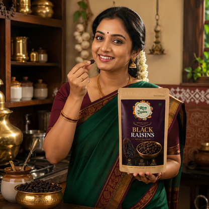 A woman in a green and maroon sari holds a bag of 'The Spice Way' Black Raisins in a warm, traditional kitchen setting.